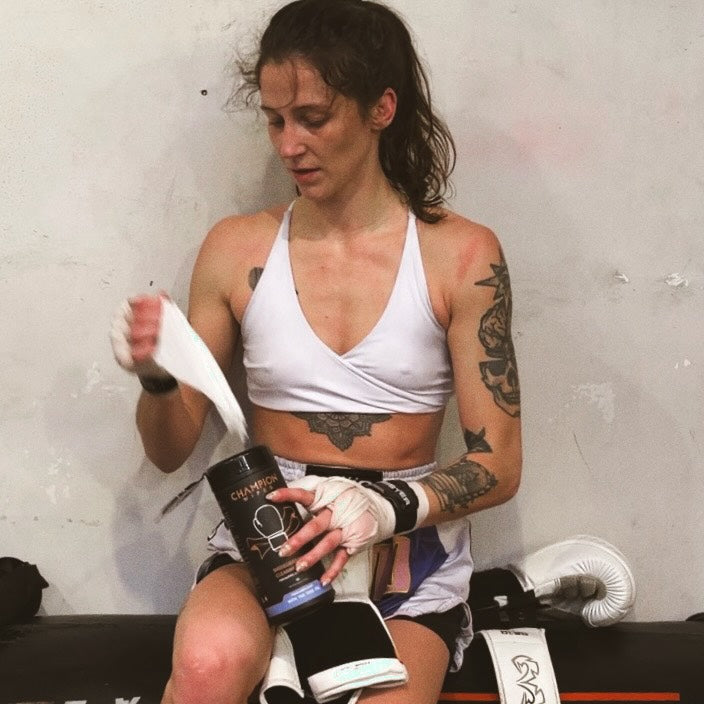 Woman preparing for a boxing session by applying tape to her hands, sitting on a black mat with red and white design.
